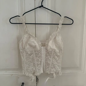 Vit spetskorsett med snörning - Superfin vit korsett med romantisk spets och snygga detaljer. Axelbanden är dekorerade med spets och toppen har snörning framtill. Croppad passform och v-ringad hals ger en trendig vibe. Perfekt för dig som vill ha en feminin och cool look.