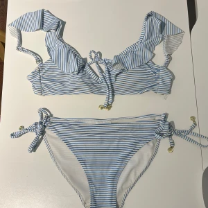 Randig bikini med volanger - Supersöt bikini med blå och vita ränder, volangdetaljer på axelbanden och snörning både upptill och nedtill. Små guldfärgade snäckor hänger på snörena, vilket är supersnyggt! 💙