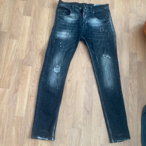 Svarta Dsquared2 jeans med slitningar - Säljer ett par svarta jeans från Dsquared2 med coola slitningar och blekta detaljer. Modellen har M passform och klassisk femficksdesign. Röd patch med 'Caten Heated' bak ger extra edge. Perfekta för dig som gillar streetstyle och vill sticka ut.