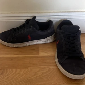 Svarta Polo Ralph Lauren sneakers - Mörk blåa sneakers från Polo Ralph Lauren med röd logga på sidan. Skorna har snörning, rund tå och vit platt sula med Polo-mönster undertill. Ovandelen är i mocka och ger en schysst streetkänsla. Perfekt för dig som gillar klassisk stil med en twist.
