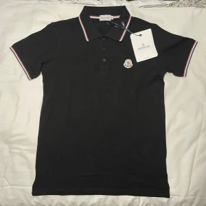 Moncler pikétröja  - Svart pikétröja från Moncler i bomull med klassisk krage och knappslå. Kragen och ärmsluten har snygga ränder i rött, vitt och blått. På bröstet sitter Moncler-loggan i broderad patch. Perfekt för dig som gillar stilrena märkesplagg.