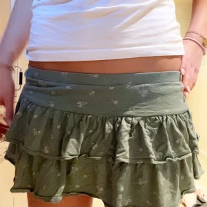 Grön blommig volangkjol - Supersöt grön kortkjol med små vita blommor och dubbla volanger. Kjolen har en bred midja och är gjord i mjukt bomullstyg som känns skönt mot huden. Perfekt för dig som gillar en chill och feminin stil. Skulle säga att den är liten i storleken så mer XS än S