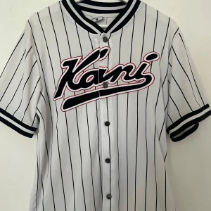 Vit randig Kani jersey med tryck - Snygg vit jersey från Kani med svarta vertikala ränder och stort broderat Kani-tryck över bröstet. Skjortan har korta ärmar, knappar framtill och sportig krage med svarta och vita ränder. Perfekt för dig som gillar streetwear och vill sticka ut.