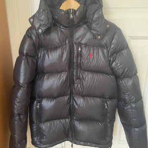 Svart dunjacka från Polo Ralph Lauren - Svart dunjacka från Polo Ralph Lauren med glansig finish och röd logga på bröstet. Jackan har en avtagbar luva, flera dragkedjeförsedda fickor och quiltad design. Perfekt för kalla dagar och är riktigt skön. Säljs för att den inte har används, 9,5/10 cond. Köpte från careofcarl för 6500:-. Skicka gärna om ni har några funderingar✌️