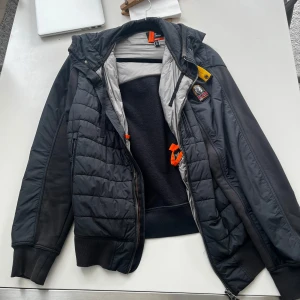 Svart pufferjacka från Parajumpers L - Svart pufferjacka från Parajumpers med coolt broderat märke på ärmen och orangea detaljer. Jackan har quiltad framsida, huva och dragkedja. Insidan är fodrad med mjuk fleece för extra värme. Perfekt för dig som vill ha en snygg och funktionell jacka med streetkänsla.       — Bara använd 10-20 gånger.