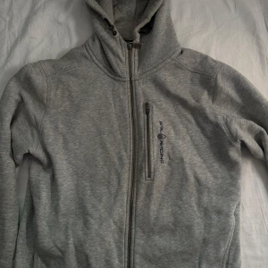 Grå hoodie från Sail Racing - Säljer en grå hoodie från Sail Racing med hel dragkedja och huva med snörning. Tröjan har en bröstficka med dragkedja och logga på framsidan. Perfekt för chill dagar och har en mjuk insida som gör den extra skön att bära.
