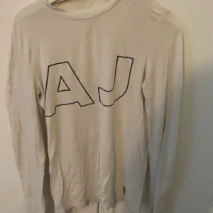Vit långärmad tröja Armani Jeans - Vit långärmad tröja från Armani Jeans med stora svarta AJ-bokstäver på bröstet. Tröjan har rund hals och är gjord i mjuk bomull. Snygg detalj med gul etikett på ärmen. Perfekt för dig som gillar stilrena och klassiska plagg med en twist.