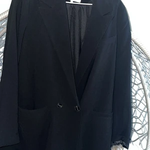 Svart klassisk kavaj med knappar - Stilren svart kavaj med två knappar framtill och klassisk krage. Rak passform och diskreta fickor på sidorna. Perfekt för dig som gillar en enkel och tidlös look. Materialet känns mjukt och har en lätt glansig finish.