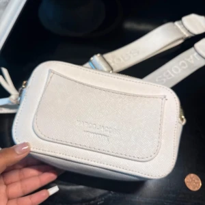 Vit axelväska från Marc Jacobs - Säljer en stilren vit axelväska från Marc Jacobs med justerbar bred rem och snygga guldfärgade detaljer. Väskan har två dragkedjor upptill och ett stort logospänne framtill. Materialet är strukturerat skinn och designen är kompakt och modern.