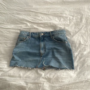 Blå jeanskjol från Zara XS - Snygg blå jeanskjol från Zara i storlek XS. Kjolen har klassisk femficksdesign, rå kant med fransar och knapp- samt dragkedja framtill. Perfekt för dig som gillar en chill och trendig look. Passformen är normal och materialet är slitstarkt denim. Orginalpris 379kr