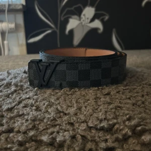 Louis Vuitton svart rutig skinnbälte - Snyggt svart bälte från Louis Vuitton med klassiskt rutigt mönster och LV-logga i metall som spänne. Bältet är gjort i skinn och har en stilren, modern look som passar till jeans eller kostym. Perfekt för dig som gillar exklusiva accessoarer.