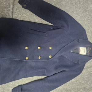 Marinblå skepparkavaj Hilfiger Denim - Säljer en marinblå skepparkavaj från Hilfiger Denim i storlek L. Jackan har klassisk dubbelknäppning med guldiga knappar, bred krage och två fickor framtill. Yttermaterialet är ull och fodret är slätt och svart med diskret logga. Perfekt för dig som gillar stilren och tidlös look.