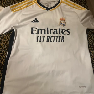 Real Madrid vit matchtröja Adidas S - Säljer en officiell Real Madrid fotbollströja från Adidas i storlek S. Tröjan är vit med svarta detaljer och gula ränder på axlarna. Broderat klubbmärke på bröstet och tryckt sponsortext. Tillverkad i lätt, ventilerande polyester med Aeroready-teknologi.