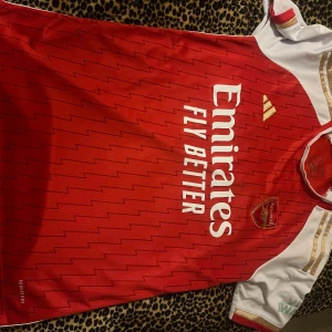 Arsenal röd kortärmad adidas tröja - Säljer en officiell Arsenal fotbollströja från adidas i storlek S. Tröjan är röd med vita och guldiga detaljer, har blixtmönster och klubbmärket broderat på bröstet. Materialet är lätt och andas, perfekt för match eller träning. Emirates Fly Better tryck på framsidan.