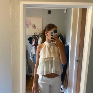 Beige volangig ärmlös blus - Säljer en superfin beige blus med volanger och hög hals. Toppen är ärmlös och har en luftig, flowy känsla med flera lager tyg som ger en romantisk vibe. Perfekt till sommaren och lätt att matcha med både jeans och kjol.