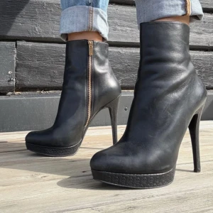 Svarta Michael Kors  stövletter med hög klack - Snygga svarta stövletter från Michael Kors i skinn med guldfärgad dragkedja på sidan. Skorna har hög klack och plattform med diskret mönster. Rund tå och stilren design som ger en cool vibe till din outfit.Michael Kors sko stl38 (24,5 cm innermått) Skinn Klack 14 cm/lätt platå  Nyskick. Sparsamt använda Vackra detaljer-gulddragkedja Ingen dålig lukt eller så.