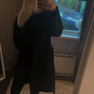 Svart kappa med bälte och stora fickor - Stilren svart kappa med stora fickor och bälte i midjan. Jackan har klassisk krage och lång ärm, perfekt för dig som gillar en clean och enkel look. Passformen är normal och materialet ser ut att vara mjukt och bekvämt.