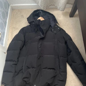 Svart pufferjacka från Canada Goose - Svart pufferjacka från Canada Goose med huva och snyggt broderat logomärke på ärmen. Jackan har dragkedja och knappar framtill, samt stora fickor. Perfekt för kalla dagar med sin tjocka och quiltade design. Stilsäker och klassisk look.