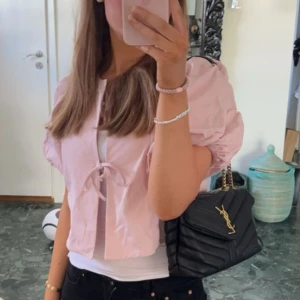 Ljusrosa blus med puffärmar från Gina Tricot - Supersöt ljusrosa blus från Gina Tricot med puffiga korta ärmar och två knyt framtill. Croppad modell som ger en trendig vibe och passar perfekt till jeans eller kjol. Tillverkad i mjuk bomull som känns skön mot huden. Perfekt för vår och sommar!