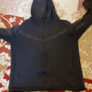 Svart hoodie från Nike med dragkedja - Svart hoodie från Nike med hel dragkedja framtill och huva. Snygga svarta detaljer över bröstet och på ärmen, samt praktiska fickor. Perfekt för chill eller träning, och tillverkad i mjuk bomullsmix för skön känsla. Storlek 160