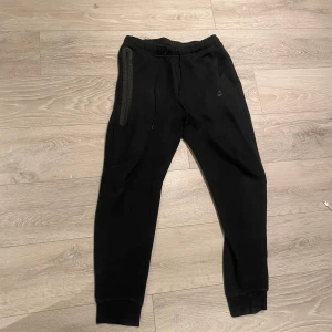 Nike tech byxor - Svarta mjukisbyxor från Nike med smal passform och ribbade muddar vid bensluten. Byxorna har snörning i midjan och en dragkedjeförsedd ficka på sidan. Perfekta för chill eller träning, med diskret Nike-logga på benet.