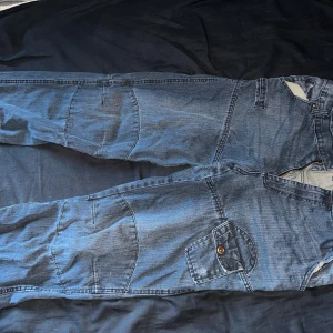 Blå Rock DNM cargojeans med fickor - Säljer ett par blå cargojeans från Rock DNM med flera stora fickor och coola sömdetaljer. Jeansen har dragkedja i gylfen och knappstängning är dock utan knapp, samt markerad midja med logga på insidan. Perfekta för dig som gillar streetwear och praktiska detaljer.