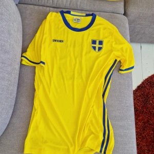 Sverige Ibrahimović 10 gul fotbollströja - Gul fotbollströja från svenska landslaget med blå detaljer, broderad text 'SWEDEN' och landslagsemblem på bröstet. Baksidan har Ibrahimović och nummer 10 i blått. Tillverkad i 100% polyester, kortärmad och med blå ränder längs sidorna och ärmarna.