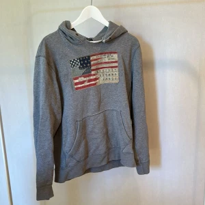 Grå hoodie Polo Jeans Company M - Säljer en grå hoodie från Polo Jeans Company i storlek M. Tröjan har en stor amerikansk flagga med vintage-tryck på bröstet och klassisk känguruficka framtill. Mjuk bomullskvalitet och huva med snörning. 