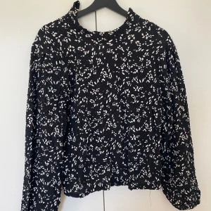 Svart  blus med lång ärm - Säljer en svart blus med vita prickar och lång ärm. Blusen har en hög krage och är gjord i ett lätt, krinklat tyg som ger en cool struktur.