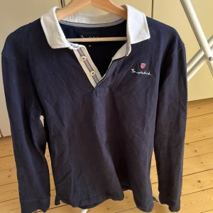 Marinblå pikétröja från Bondelid - Snygg marinblå pikétröja från Bondelid med vit krage och knappslå. Tröjan har lång ärm och broderad logga på bröstet. Perfekt för dig som gillar klassisk stil med sportig touch. Materialet är mjuk bomull som känns skönt mot huden.