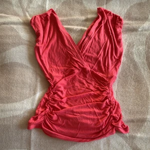 Röd omlott topp med v-ringning - Snygg mörkrosa omlott topp med djup v-ringning och rynkade sidor som ger en cool och figurnära siluett. Toppen är ärmlös och gjord i mjukt bomullsmaterial, perfekt för att lyfta din outfit med lite färg.