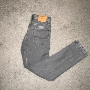 Grå loose fit jeans från Jack & Jones - Säljer ett par grå jeans från Jack & Jones i loose fit-modell. De har klassisk femficksdesign, raka ben och en snygg tvättad look. Jeansen är tillverkade i slitstarkt denim och har en läderpatch med logga baktill i midjan. Perfekt för dig som gillar avslappnad stil. Riktigt sparsamt använda så skicket är 9/10