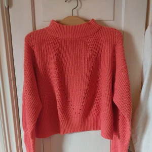 Stickad korallrosa tröja  - Säljer en stickad tröja i en snygg korallrosa/laxrosa färg med hög ribbad krage och långa ärmar. Tröjan har ett coolt triangulärt hålmönster på framsidan och på ärmarna, men hålen är så pass små att man inte tvunget måste ha en tröja under. Materialet är i akryl så det är inte det minsta stiksigt mot hunden så som ull kan vara.