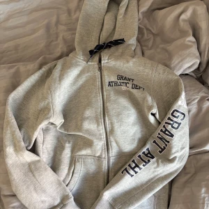 Grå hoodie från Grant Athletics - Snygg ljusgrå hoodie från Grant Athletics med marinblå tryck på bröstet och ärmen. Tröjan har huva med mörkblå snörning, dragkedja framtill och mjukt fleecefoder. Knappt använd!