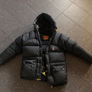 Svart dunjacka från Fjällräven XXXS -  (PRIS KAN DISKUTERAS VID SNABB AFFÄR) Säljer en svart dunjacka från Fjällräven i storlek XXXS. Inte smutsig alls. Jackan är inköpt i April, endast använd nårgra fåtal gånger under påskveckan 2025. Den har ett litet litet brännhål men jag har satt en för stor tejp (går att ta bort och sätta en finare svart tejp” Jackan är från fjällrävens hemsida och är i storlek XXXS men sitter som en XS-S