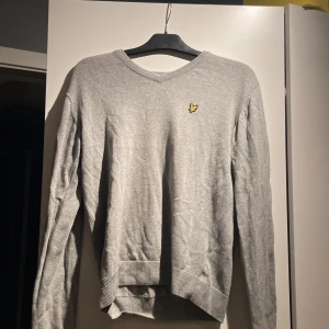 Grå stickad tröja Lyle & Scott - Snygg grå stickad tröja från Lyle & Scott med klassisk gul broderad fågel på bröstet. Tröjan har v-ringad hals och ribbade muddar vid ärmslut och nederkant. Perfekt för dig som gillar stilrena och enkla plagg med en ikonisk detalj.