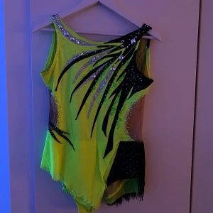 Neongul glittrig dansklänning med fransar - Säljer en unik neongul dansklänning med svarta detaljer och glittrande paljetter. Klänningen har asymmetrisk design, meshpartier och fransar på ena sidan. Den är ärmlös och har en cool, edgy look som verkligen sticker ut på dansgolvet.                       Nypris för en liknande dräkt ligger på drygt 2500 kr, men priset kan diskuteras vid snabb affär. Den är i bra skick och har bara använts några gånger. Storleken är ungefär S/M.