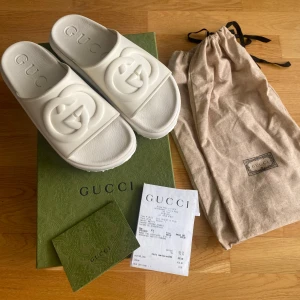 Gucci sandaler - Snygga vita sandaler från Gucci med platåsula och tydlig logga framtill. Tillverkade i syntetmaterial med präglad GUCCI-text i fotbädden. Perfekta för dig som vill sticka ut med en clean och lyxig vibe. Kommer med originalpåse och box. Använda endast 1 gång