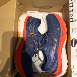 Blåa Asics sneakers med röd sula - Snygga Asics sneakers i marinblått med röda och gula detaljer. Skorna har en sportig design med snörning och en färgglad sula i rött, vitt och gult. Perfekta för dig som gillar att sticka ut och vill ha bekväma skor till träning eller vardag.