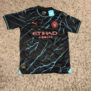 Manchester City tröja - Snygg Manchester City tröja från Puma i svart med ljusblå blixtmönster och rosa detaljer. Kortärmad modell med klubbmärke och Etihad Airways-logga på bröstet. Helt oanvänd med tags kvar. Passar perfekt för sommaren.