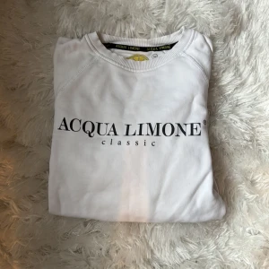 Vit sweatshirt från Acqua Limone - Vit sweatshirt från Acqua Limone med klassisk svart logga framtill och stor gul citron-grafik på ryggen. Tröjan har rund hals, långa ärmar och ribbade muddar. Perfekt för dig som gillar stilrena och fräscha plagg med en twist.