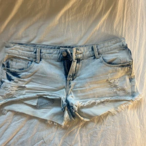Lågmidjade slitna jeansshorts - Snygga ljusblå jeansshorts med slitna detaljer och fransig kant. Klassisk femficksmodell med låg midja och dragkedja. Perfekta för varma dagar och ger en avslappnad vibe. Materialet är mjukt denim och shortsen har en cool, tvättad look.
