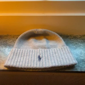 Grå stickad mössa från Polo Ralph Lauren - Snygg grå mössa från Polo Ralph Lauren med klassisk ribbad kant och broderad blå logga framtill. Tillverkad i mjuk ull som håller dig varm under kyliga dagar. Perfekt accessoar för att lyfta din vinterstil kom med bud 🤌