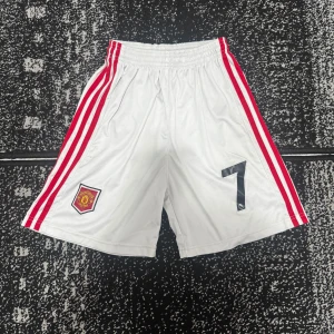 Manchester United vita fotbollsshorts - Vita Manchester United shorts med röda ränder på sidorna, broderad klubbmärke och svart nummer 7. Elastisk midja med snörning för perfekt passform. Tillverkade i lätt polyester, perfekta för fotbollsträning eller match.