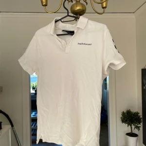 Pikétröja  - Vit piké från Peak Performance i bomull, klassisk krage med knappar och svart logga på bröstet. Snygg svart detalj på ena ärmen. Perfekt för en clean och sportig stil. STLK M.