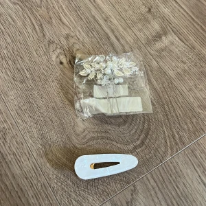 Hårspänne och hårdekoration med pärlor - Säljer ett vitt hårspänne i skimrande plast och en elegant hårdekoration med silverfärgade blad och vita pärlor. Perfekt för att ge frisyren en extra touch med glittriga detaljer och romantisk känsla.
