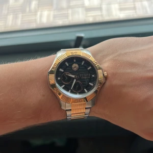 Versus Versace klocka i guld och silver - Snygg klocka från Versus Versace med svart urtavla, guld- och silverfärgad boett och länkarmband. Klockan har detaljer i guld runt urtavlan och på länken, samt Versus-logga och lejonmotiv på urtavlan. Perfekt accessoar för dig som gillar lyxig stil.