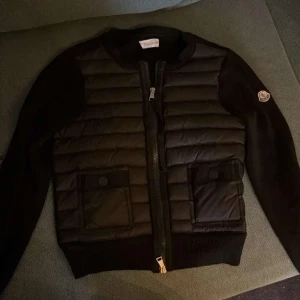 Svart cardigan från Moncler - Säljer en svart pufferjacka från Moncler med quiltad front och två stora fickor med knappstängning. Jackan har långärmade stickade ärmar och Moncler-logga på ena ärmen. Dragkedja framtill och stilren, sportig look. Perfekt för dig som vill ha något snyggt och bekvämt.