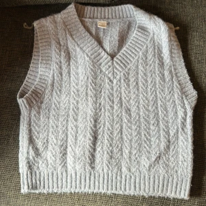 Ljusgrå stickad väst från Lindex - Mysig ljusgrå stickad väst från Lindex med kabelstickat mönster och bred ribbad kant vid hals, ärmhål och nederkant. Perfekt att slänga över en skjorta eller t-shirt för en chill och trendig look.