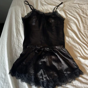 Svart linnetopp med spetsdetaljer - Säljer en svart linnetopp med tunna axelband och snygga spetsdetaljer längs urringningen. Toppen är i glansigt material och har en feminin, elegant look. Perfekt att matcha med kjol eller jeans för en trendig vibe.